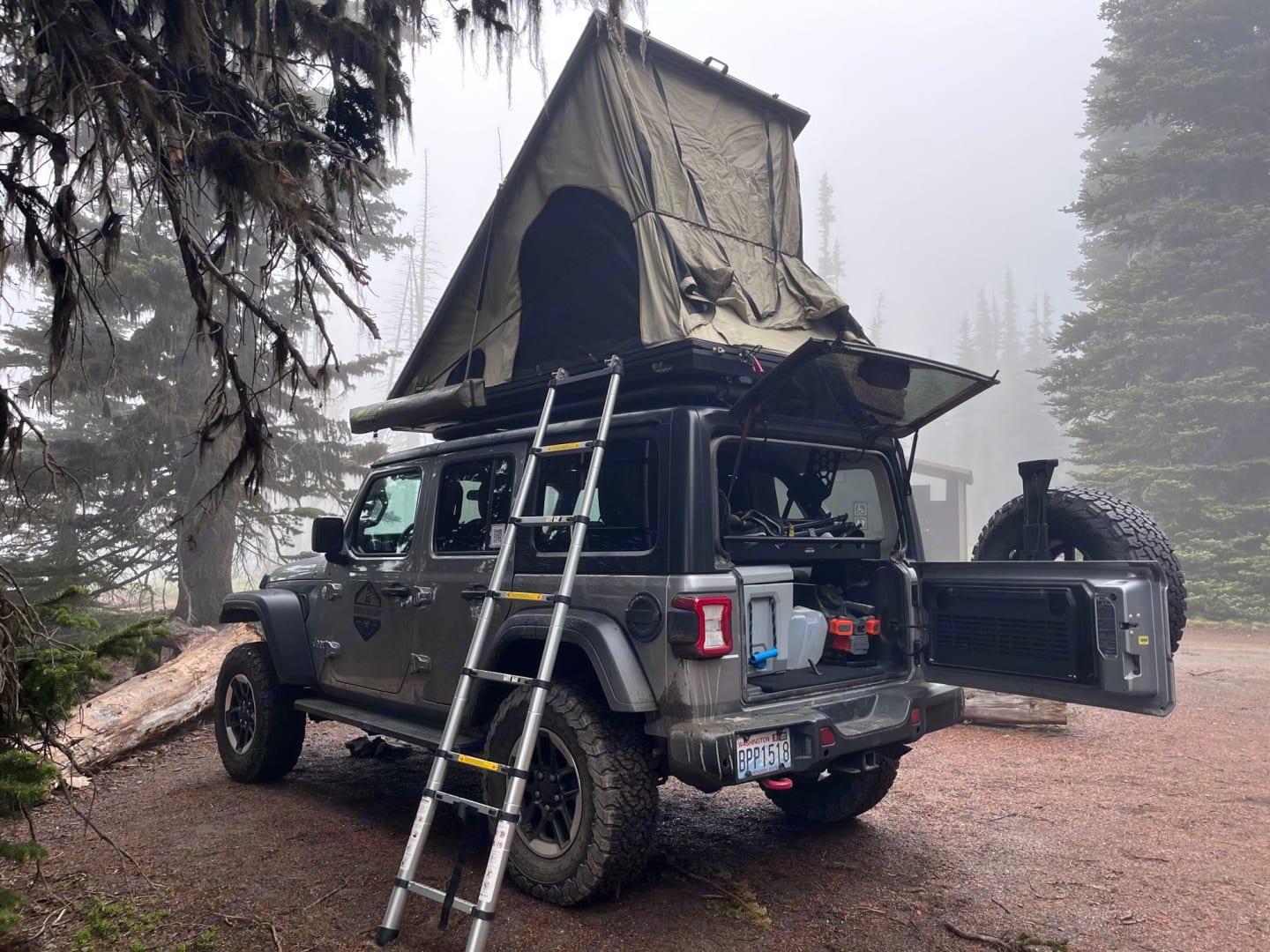 A Jeep Gladiator with rooftop tent set up and tailgate open at a foggy Pacific Northwest campsite